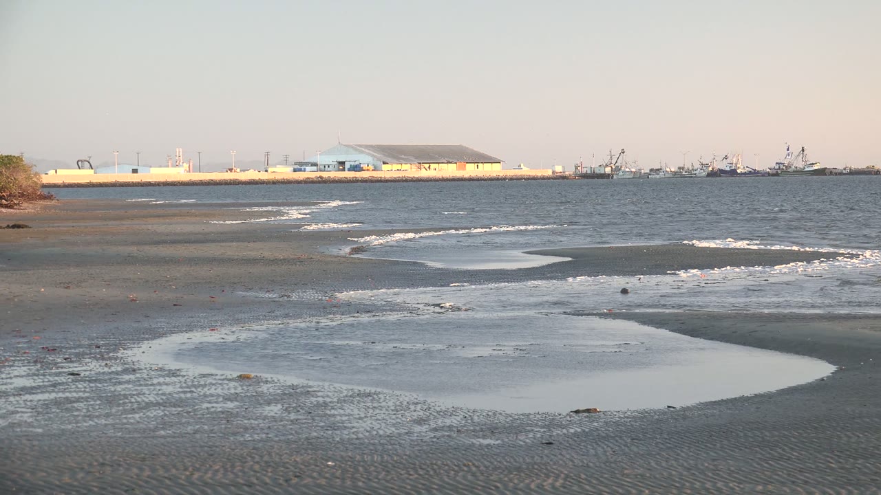 At 0:02 in San Carlos, Baja California Sur, a broad shallow pool reflects the pale sky as small waves break behind it near distant waterfront buildings and boats, with no visible people.