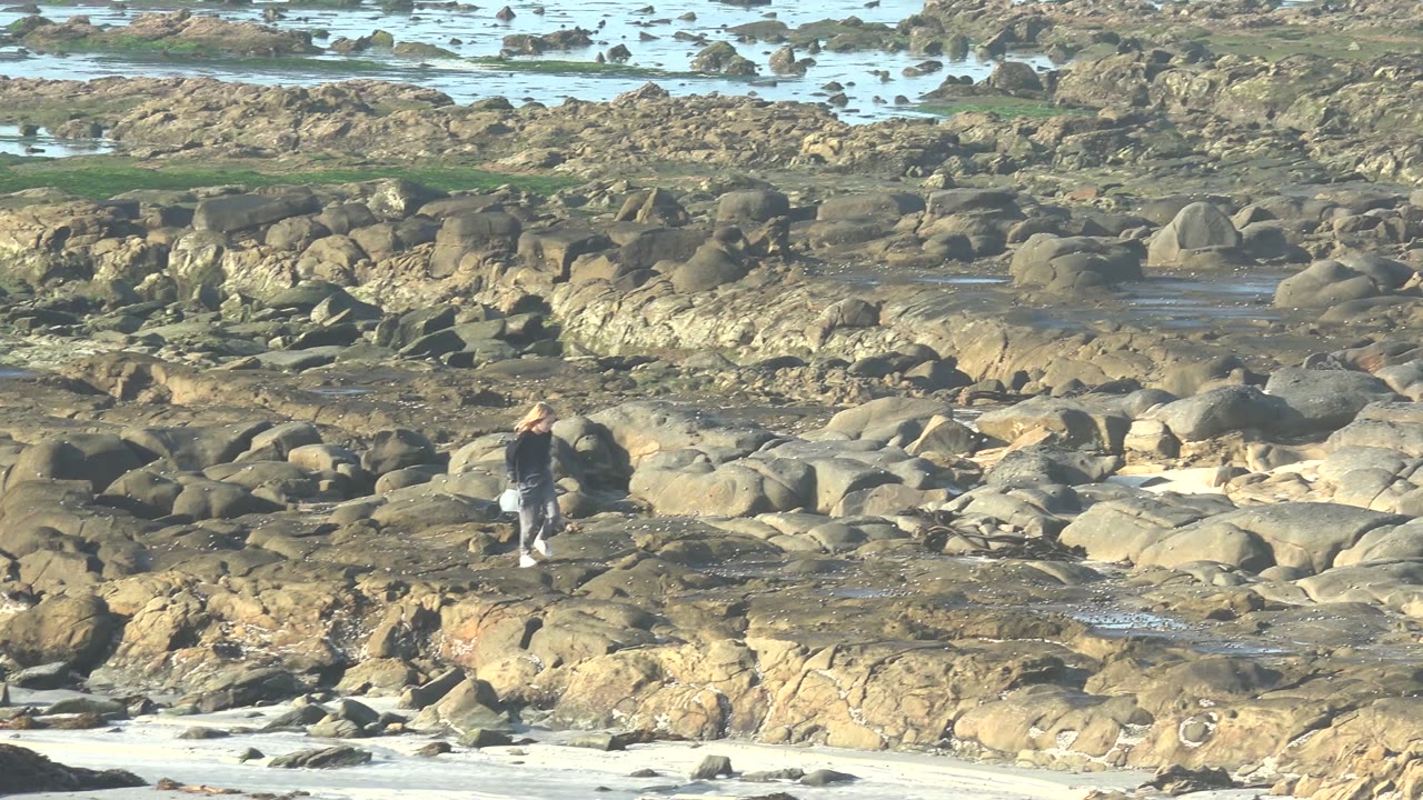 A person walks across wet rock shelves on the La Bocana shoreline, with pale surf and scattered coastal rocks in the background.