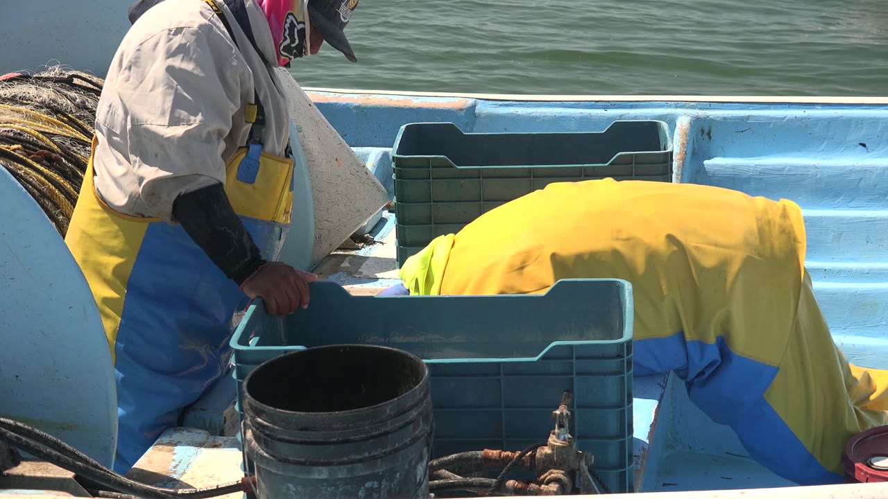 Two fishermen in yellow and blue rain gear work over a blue crate on a small boat in La Bocana, with green ocean water behind them.