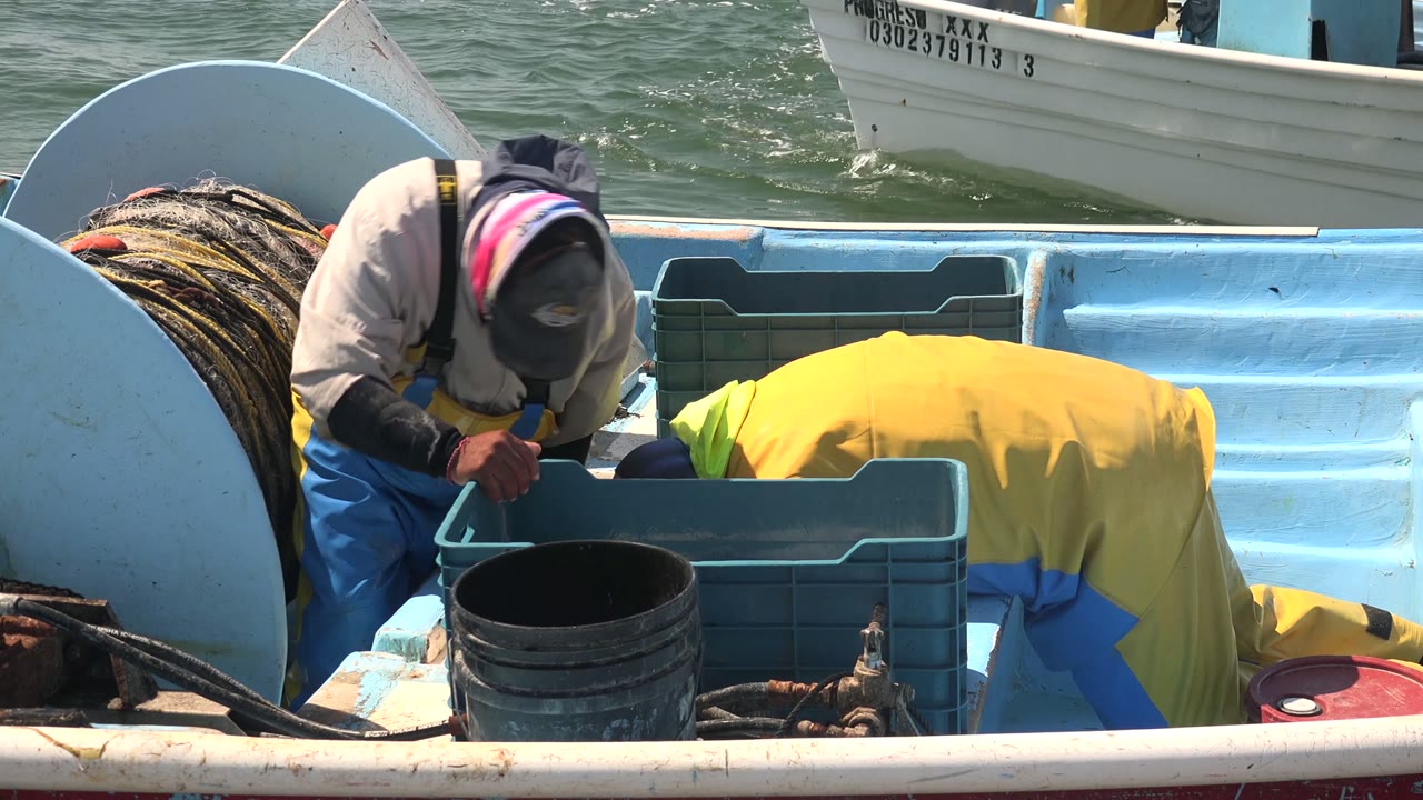 At 0:00 in La Bocana, Baja California Sur, two fishermen in yellow and blue rain gear lean over crates on a light blue boat deck with ocean water behind them.