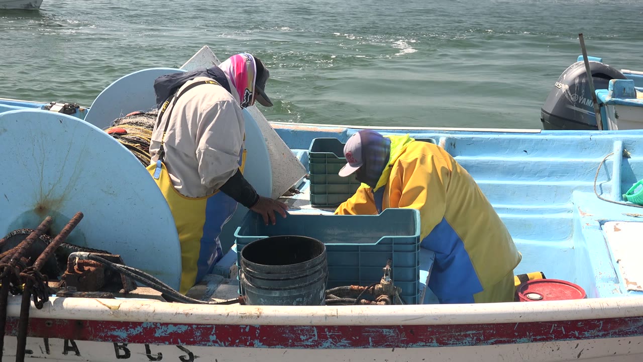 Two fishermen in hats and rain gear sort catch in plastic crates on a small blue boat in La Bocana, Baja California Sur, with fishing gear in the foreground and ocean water behind them.