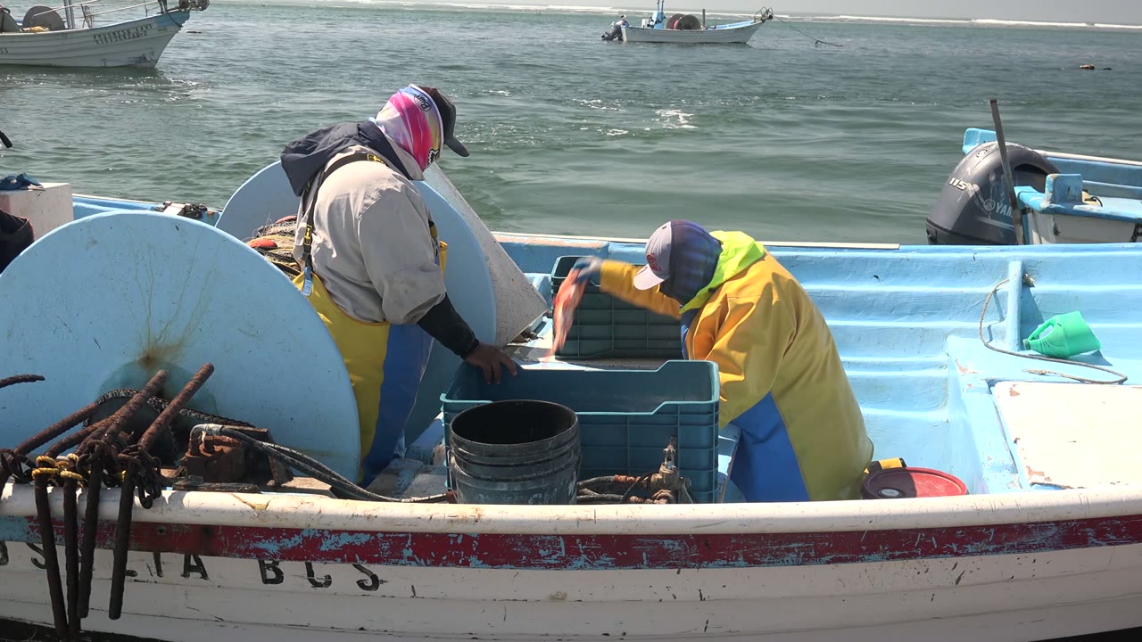 Two fishermen continue working beside blue plastic crates on the small boat in La Bocana, while the ocean surface and distant boats fill the background.