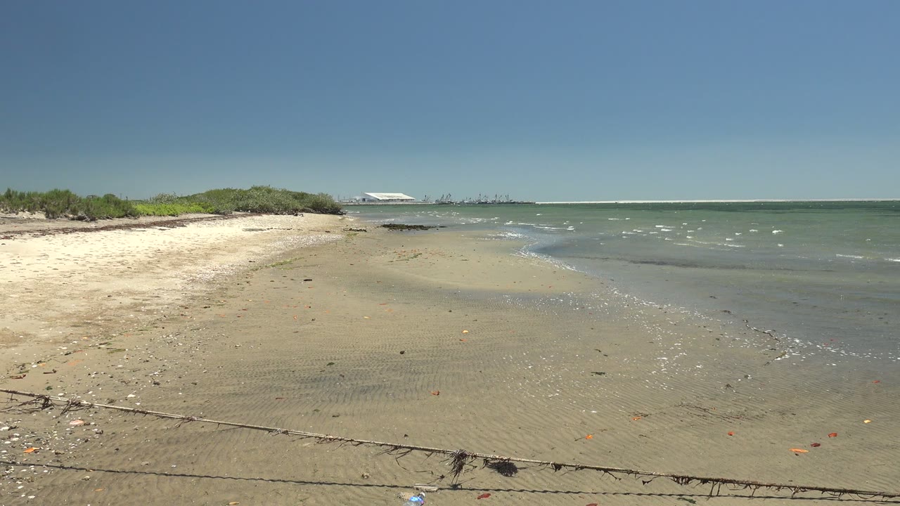 At 0:02 in San Carlos, Baja California Sur, the pale sandbar and shallow green-blue water fill the frame as small whitecaps appear offshore and the distant structure sits left of center, with no visible people.