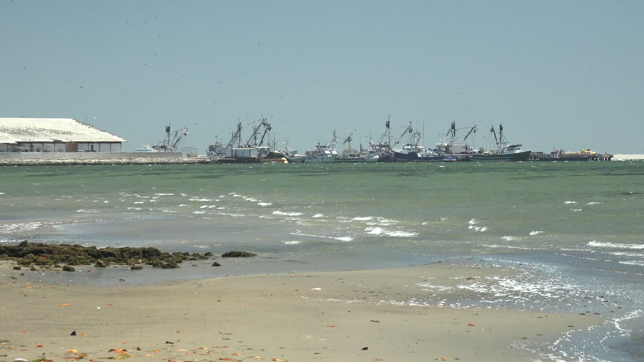 Shallow surf curves across sand with rocks and port cranes in the distance.