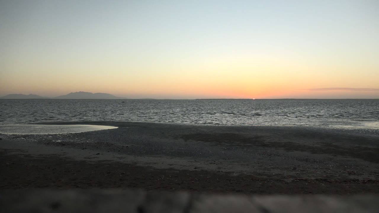 At 0:00 in San Carlos, Baja California Sur, wet sand leads to calm rippled ocean, distant mountain silhouettes, and an orange horizon under pale blue sky with no visible people.