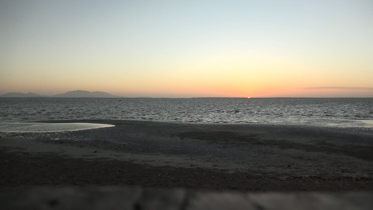 San Carlos, Baja California Sur beach at sunset with dark wet sand, a small reflective tide pool, rippled ocean, distant islands on the horizon, and no visible people.