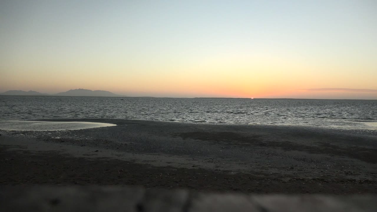 San Carlos, Baja California Sur dusk beach view with a pale tide pool on dark sand, calm ocean bands, island silhouettes in the distance, and no visible people.