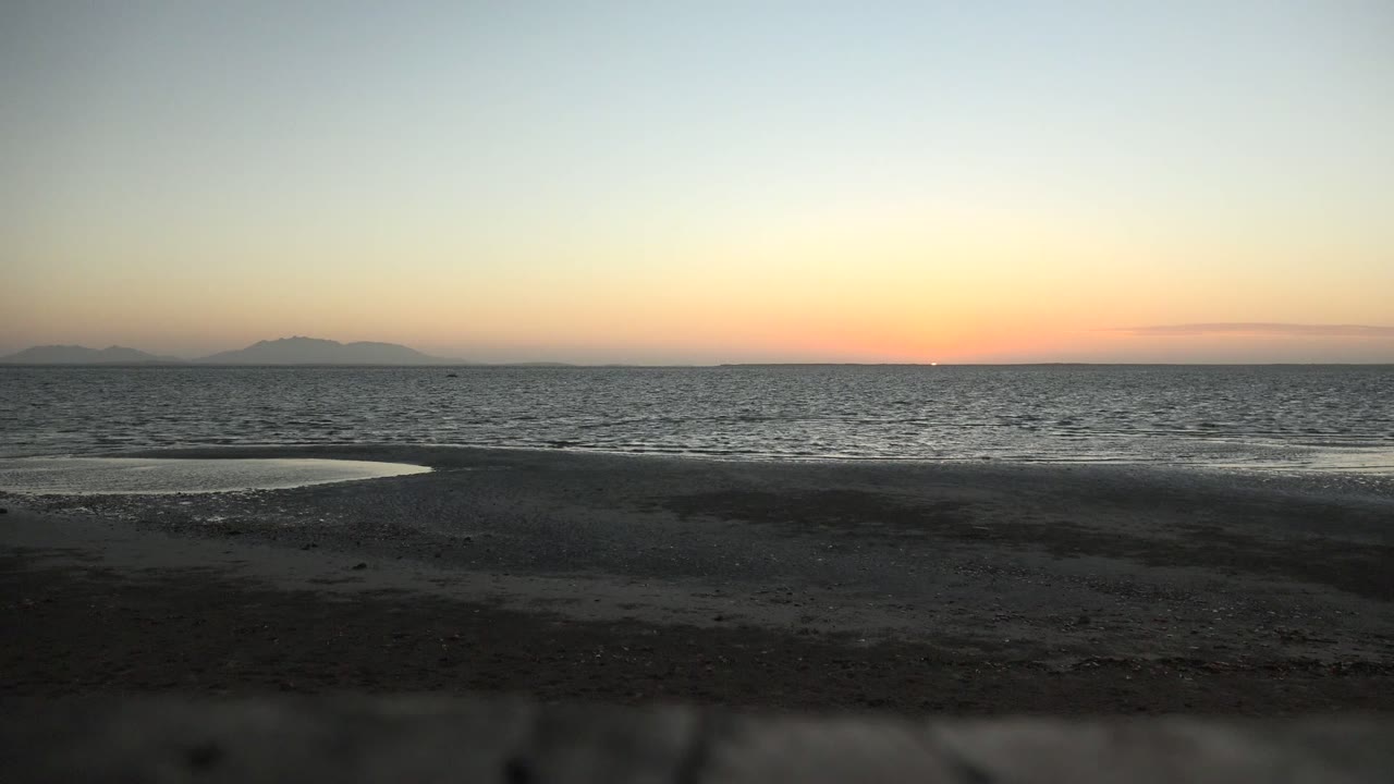 Wide sunset view of San Carlos, Baja California Sur with dark wet sand, a shallow shore puddle at left, calm ocean, distant mountain silhouettes, and no visible people.