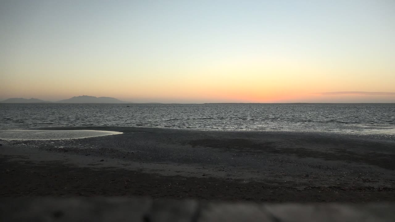 San Carlos, Baja California Sur beach at dusk with dark sand in front, rippled ocean beyond, faint island silhouettes on the horizon, and no visible people.