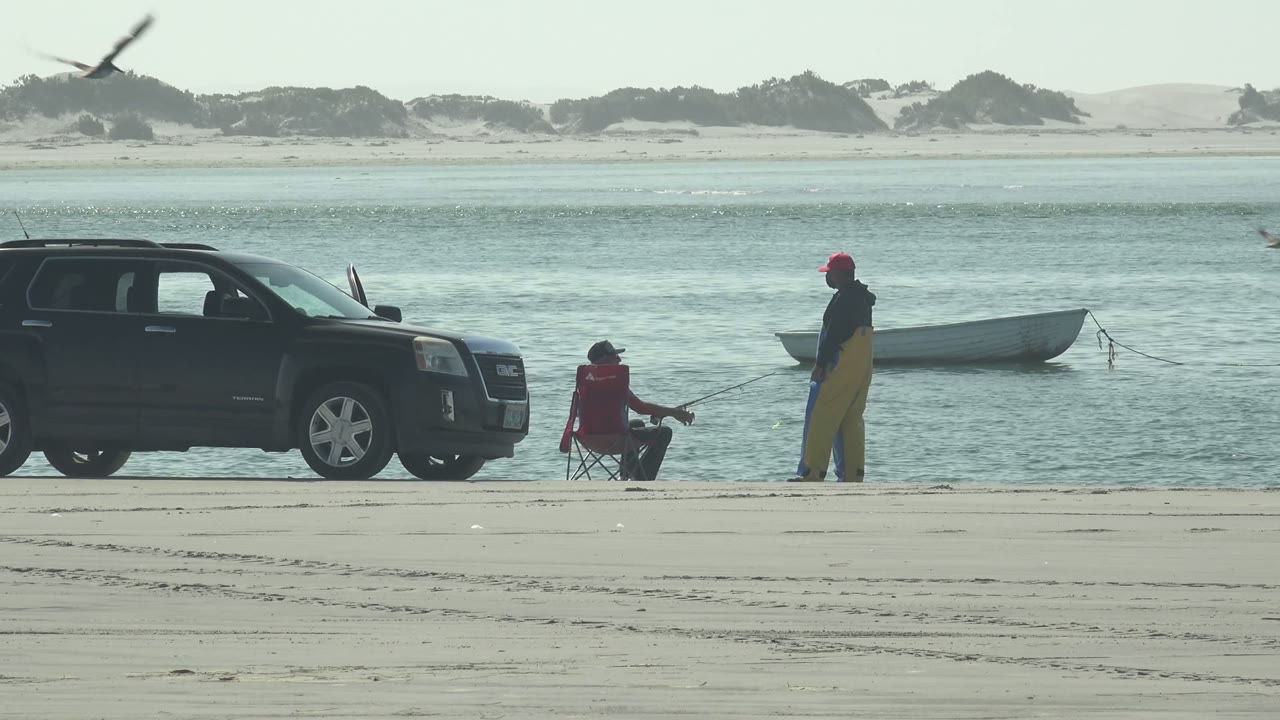 At La Bocana, Baja California Sur, two people fish from the sandy shore beside a black SUV while a small white boat floats in the water behind them and a bird crosses the upper left frame.
