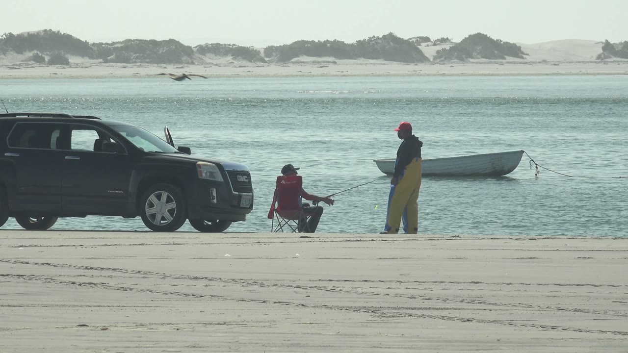 A seated person and a standing person fish near a black SUV with a small boat on the water.