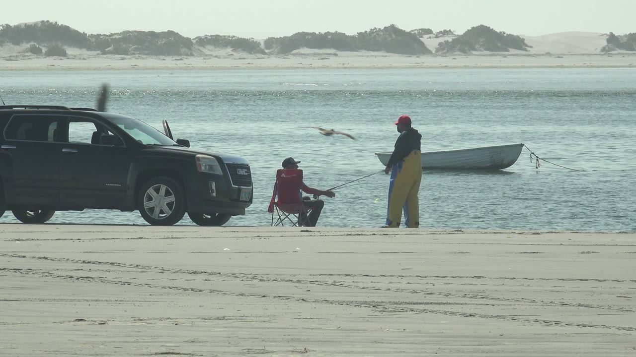 At La Bocana, Baja California Sur, the black SUV sits just left of the two fishers, the small boat rests on the water behind them, and birds pass across the open shoreline view.