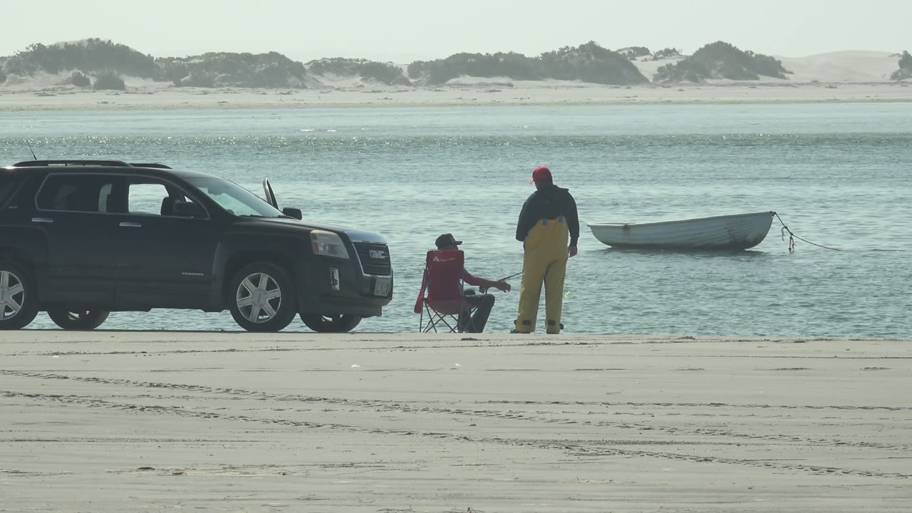 Opening frame in La Bocana, Baja California Sur, with a black SUV at left, one person seated in a red chair, another standing in yellow overalls, and a small boat floating near the shore.