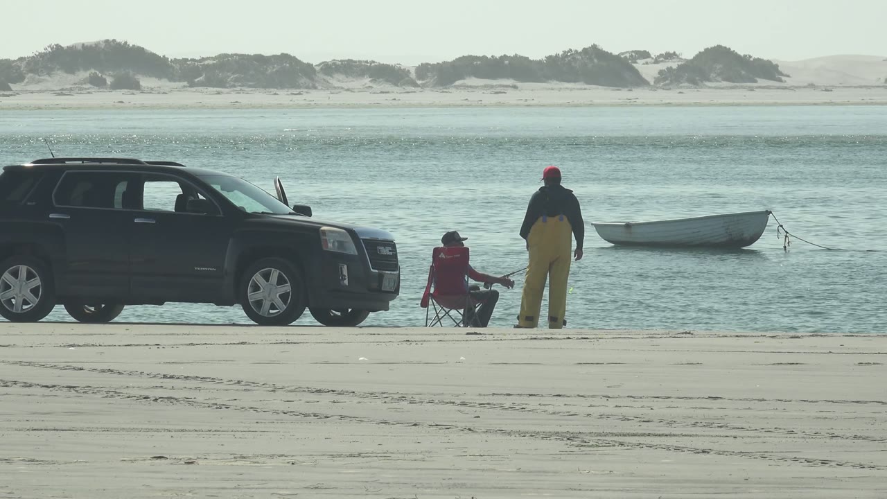 Beach scene with black SUV, seated person, standing person, and small boat offshore.