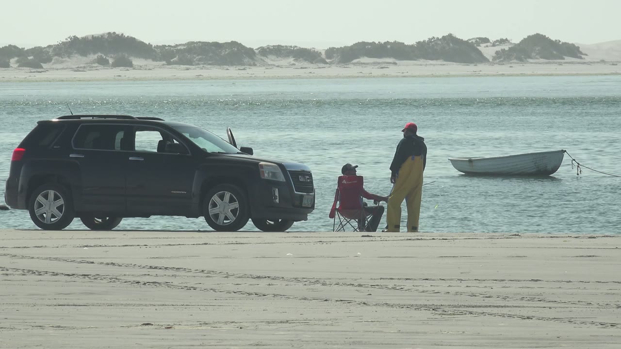 At 0:00 in La Bocana, Baja California Sur, a black SUV is parked on the sandy shore while one person sits in a red chair and another stands near a small boat on calm water.