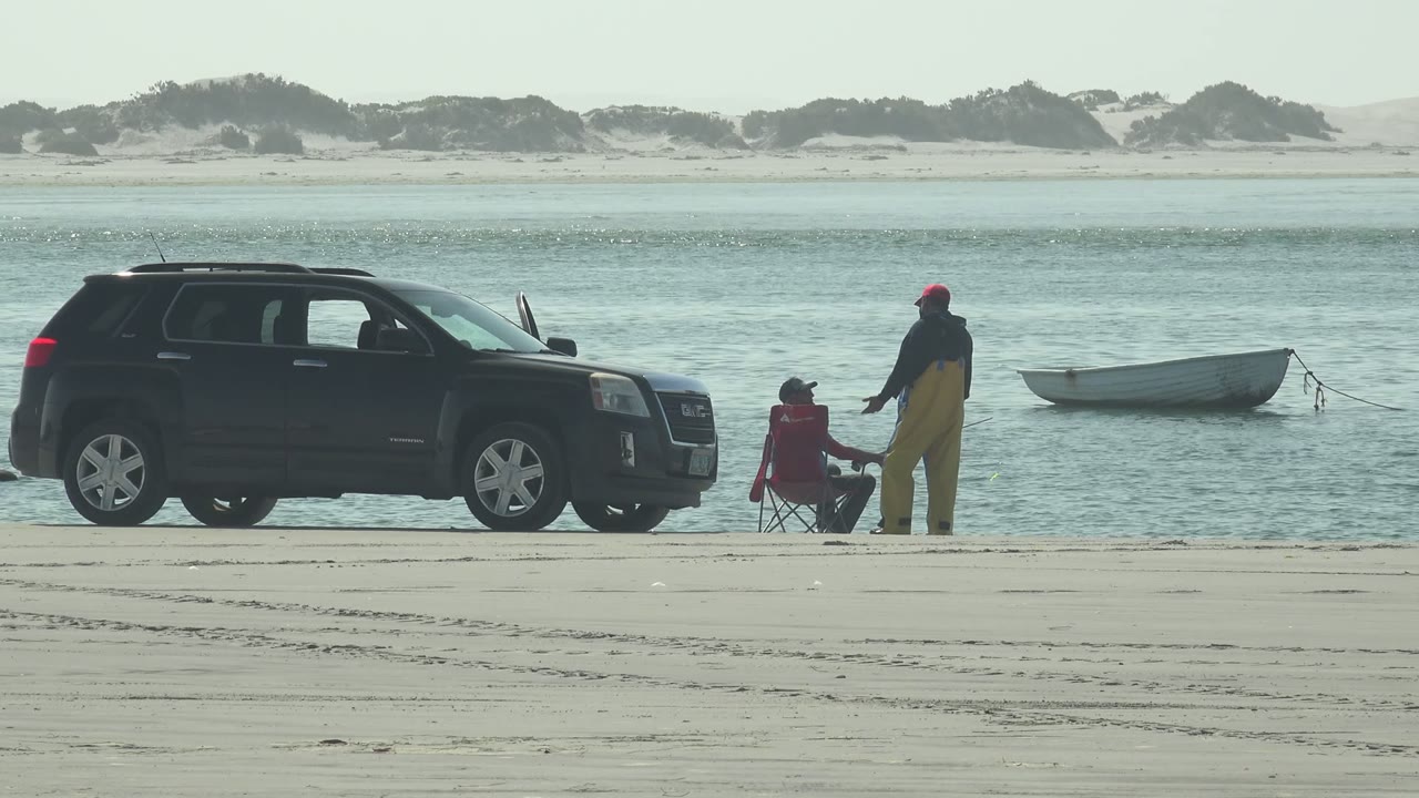 Standing beachgoer near a seated person as a small boat floats behind them.