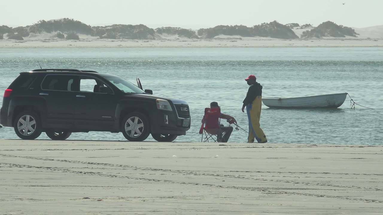 At La Bocana in Baja California Sur, two visible people fish near a dark SUV, a red folding chair, and a small white boat on calm water.