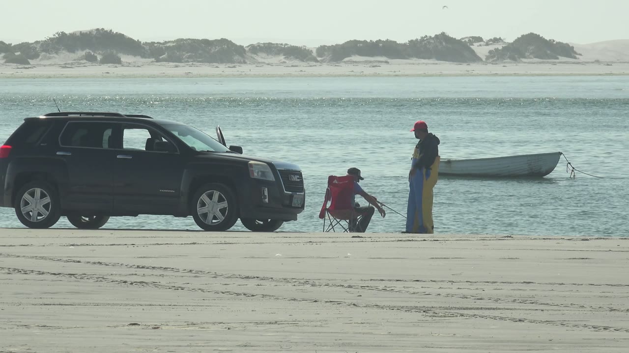 Standing angler and seated angler fish near a parked SUV and moored boat.