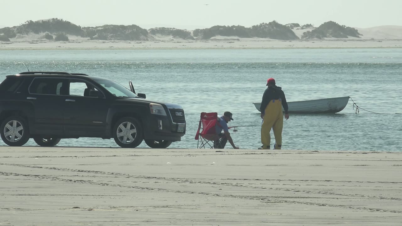 At La Bocana, the seated person reaches toward fishing gear by the red chair while the standing person remains near the water with the SUV and boat framing the scene.