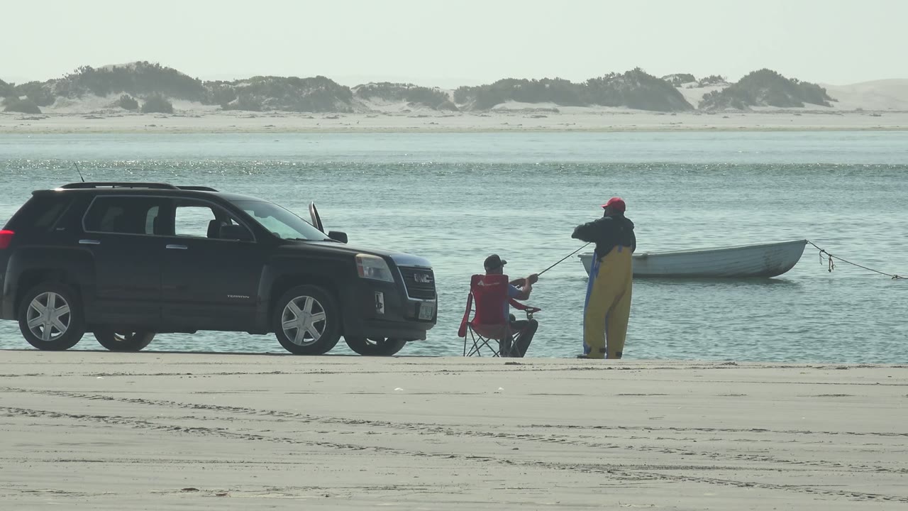 At 0:00 in La Bocana, Baja California Sur, a seated person in red and a standing person in yellow waders fish beside a dark SUV with a small boat floating behind them.
