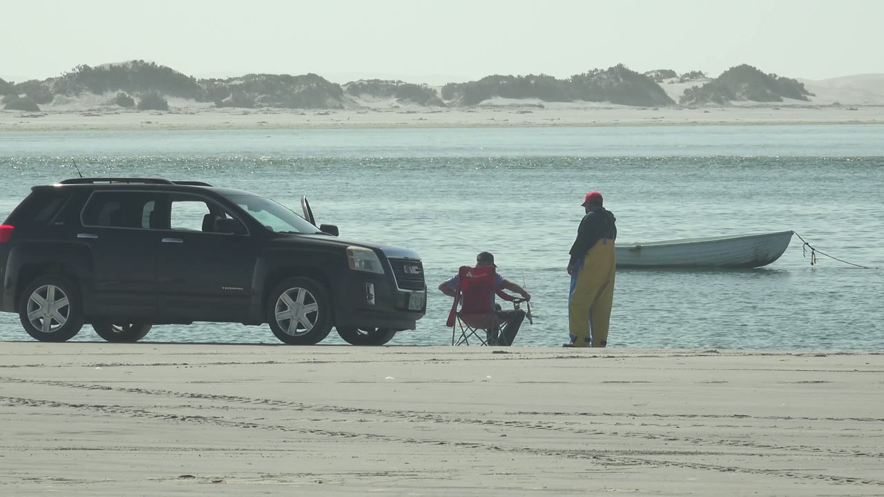 At 0:02 in La Bocana, Baja California Sur, the seated person in red raises the fishing rod toward the standing person, with the SUV on the left and a pale boat lined up behind them on the water.