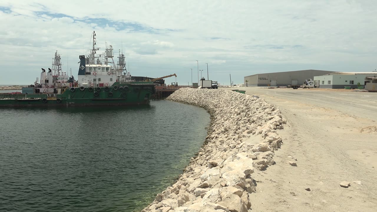 Opening frame of a green and white tugboat beside a pale rocky breakwater in Guerrero Negro, Baja California Sur, with calm water, industrial buildings, and no visible people.