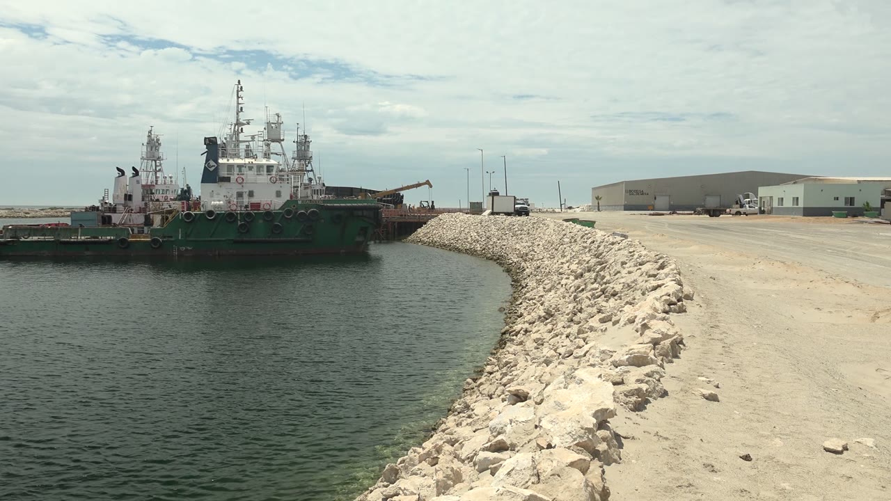 Green and white tugboat mid-frame on calm harbor water in Guerrero Negro, Baja California Sur, with the curved rock breakwater and low industrial buildings behind it, no visible people.