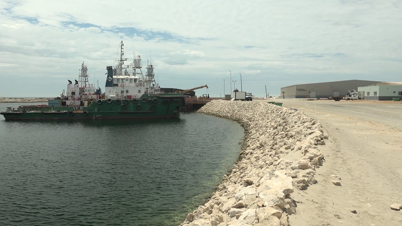 Tugboat crossing the midground beside the pale stone breakwater in Guerrero Negro, Baja California Sur, with a dirt road, warehouse-like buildings, and no visible people.