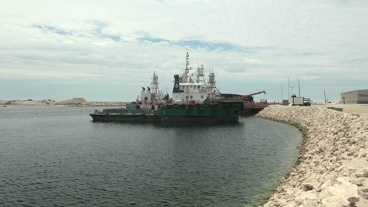 A green and white work vessel floats near a pale rock breakwater in Guerrero Negro, Baja California Sur, with sand dunes and a cloudy sky behind it and no visible people.