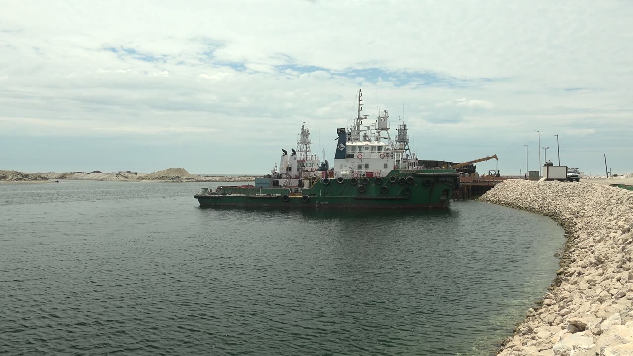 A large green-hulled ship sits just off the rocky edge of the waterway in Guerrero Negro, with small ripples in the foreground, pale dunes in the distance, and no visible people.