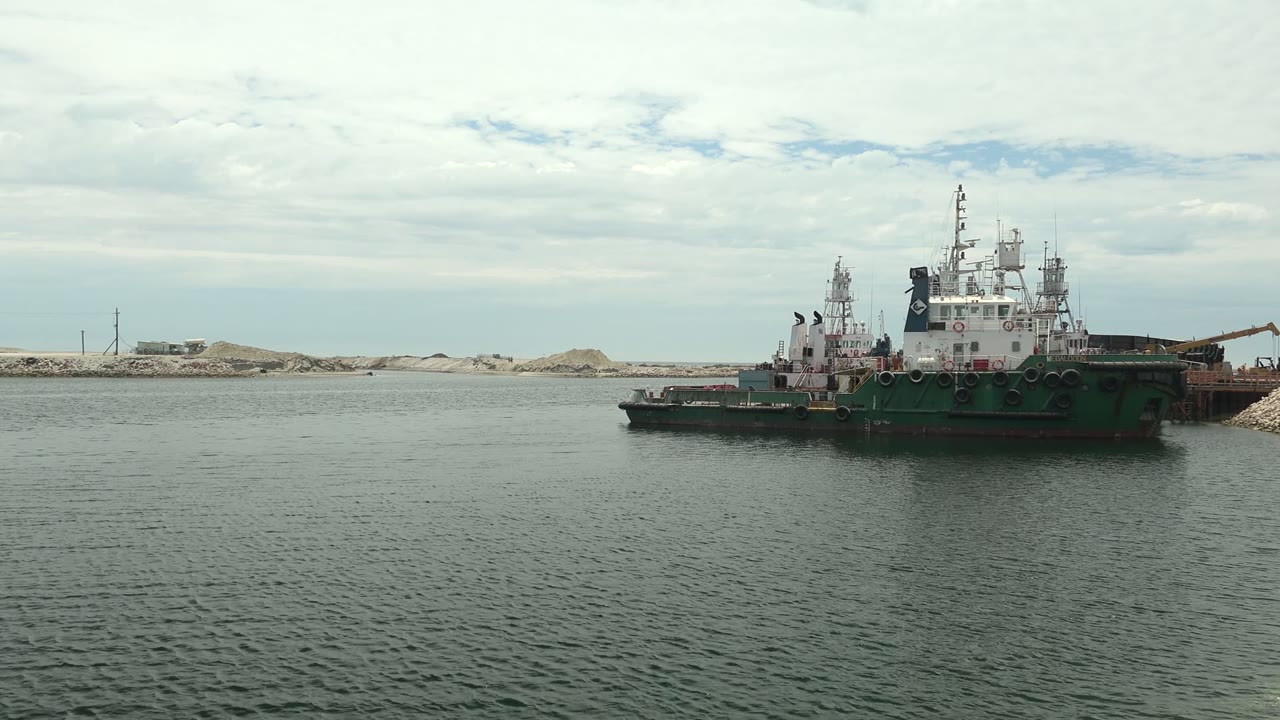 At 0:00, a green and white tugboat sits midframe on calm water in Guerrero Negro, Baja California Sur, with sandy shore and low industrial structures behind it and no visible people.