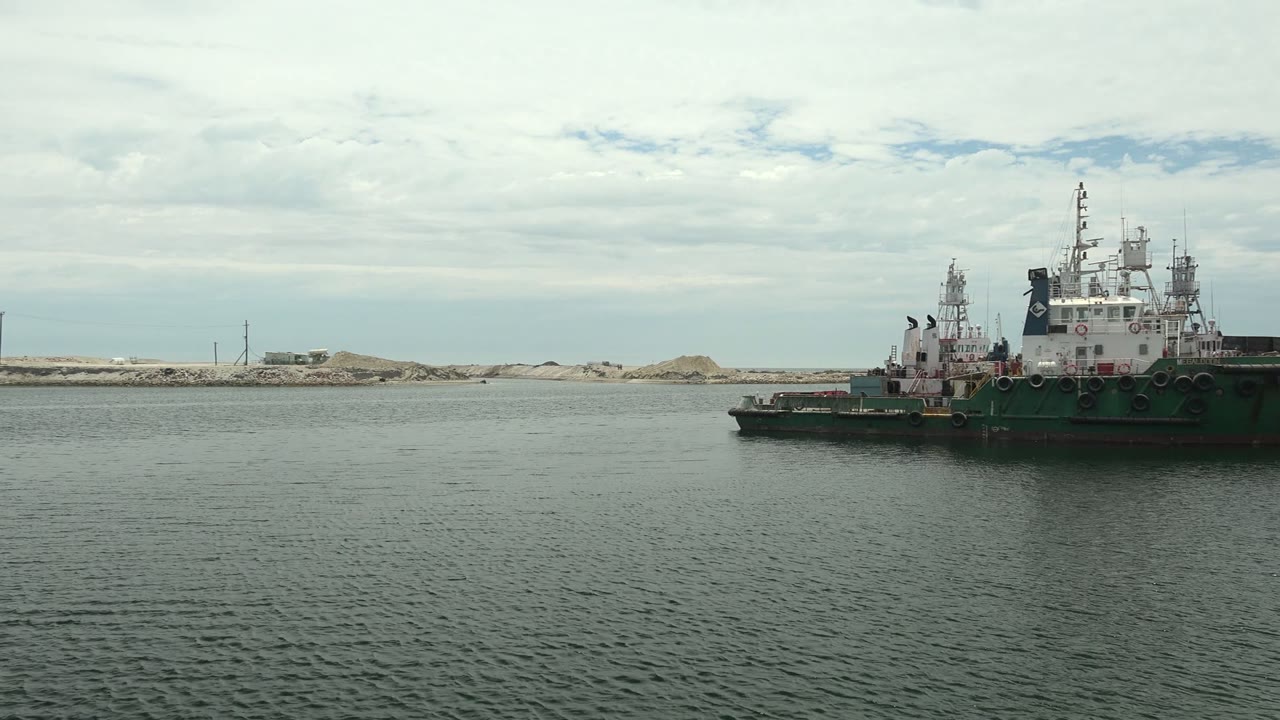 At 0:02, the tugboat is pushed toward the right side of the frame in Guerrero Negro, leaving open rippled water across the foreground and a sandy industrial shore behind it, with no visible people.
