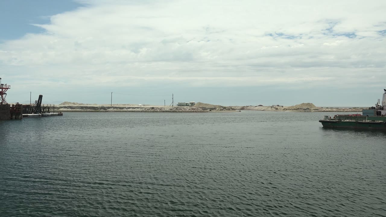 Calm harbor water in Guerrero Negro with the edge of an industrial dock and crane structure just entering from the left, sandy shoreline and cloudy sky beyond, no visible people.