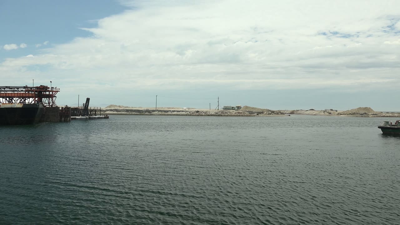A crane-topped dock structure is centered over rippled harbor water in Guerrero Negro, with pale sand piles and distant utility poles beneath a partly cloudy sky, no visible people.
