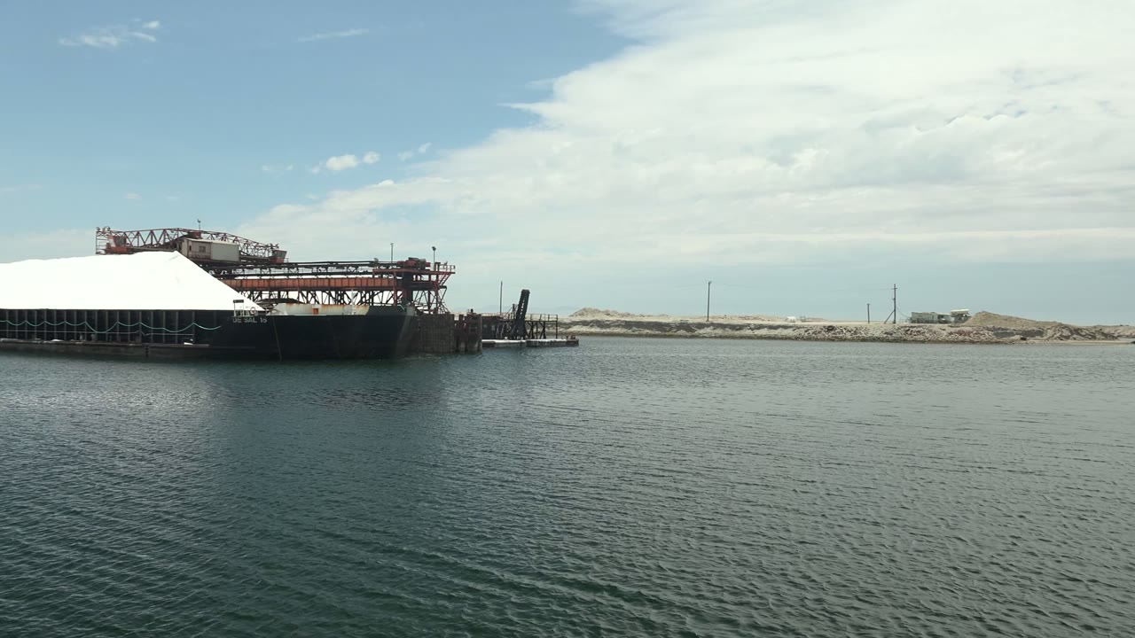 At 0:00 in Guerrero Negro, a large covered barge sits left of center across calm water with dock equipment, pale shoreline, clouded sky, and no visible people.