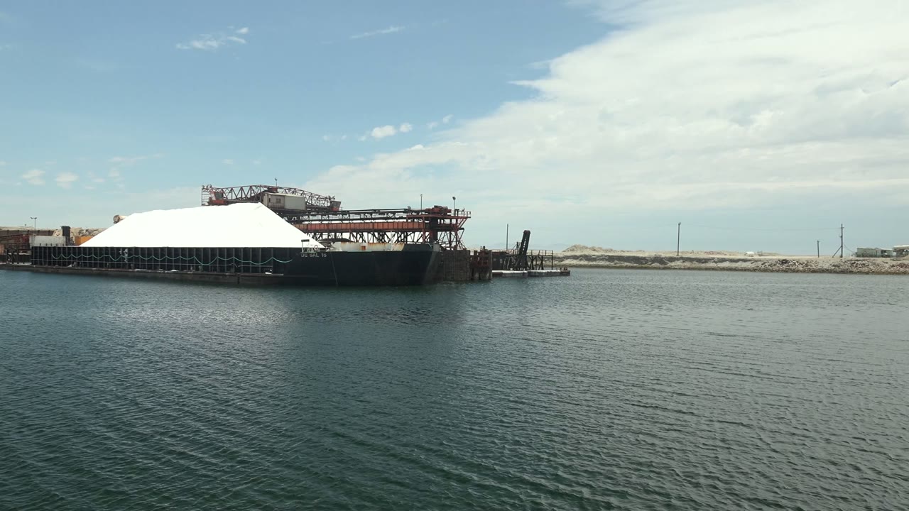 At 0:02 in Guerrero Negro, the barge remains near center with its white cover bright against darker hull supports, calm water below, distant land behind, and no visible people.