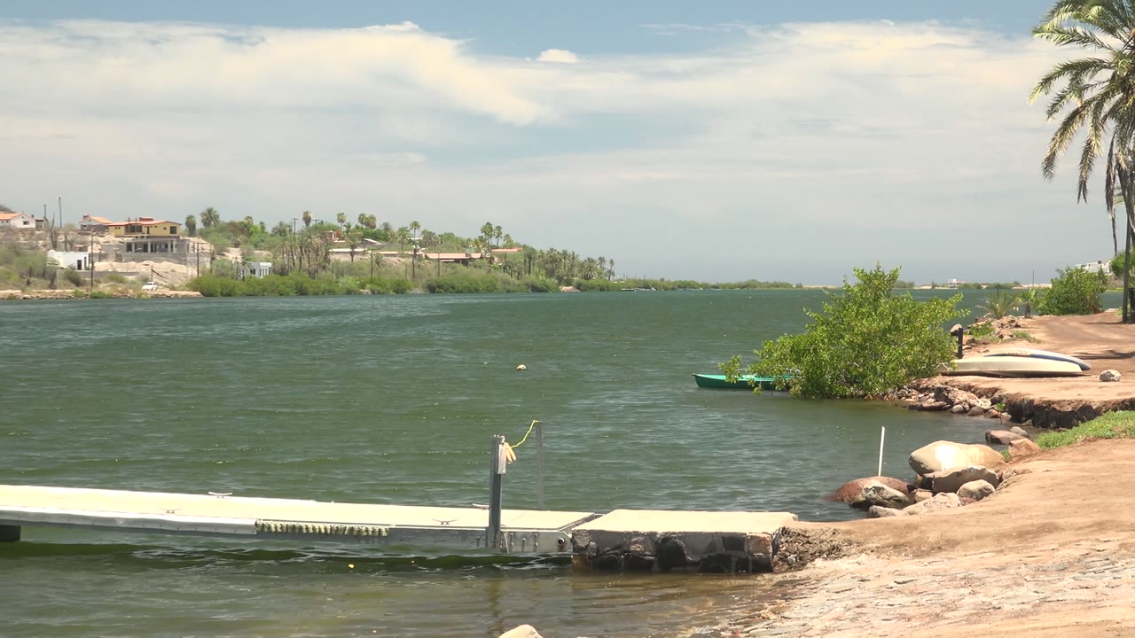 At 0:01, the Mulege waterfront remains framed with the dock across the lower edge, a small boat near the right shore, distant houses, and no visible people.