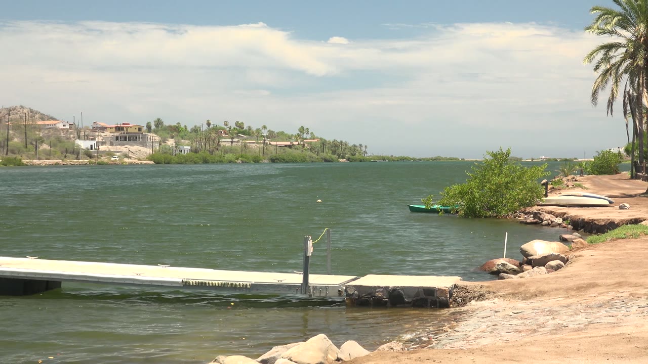 At 0:02, light ripples cross the water around the dock while the sandy Mulege shoreline, palms, rocks, small boat, and distant homes stay visible with no people seen.