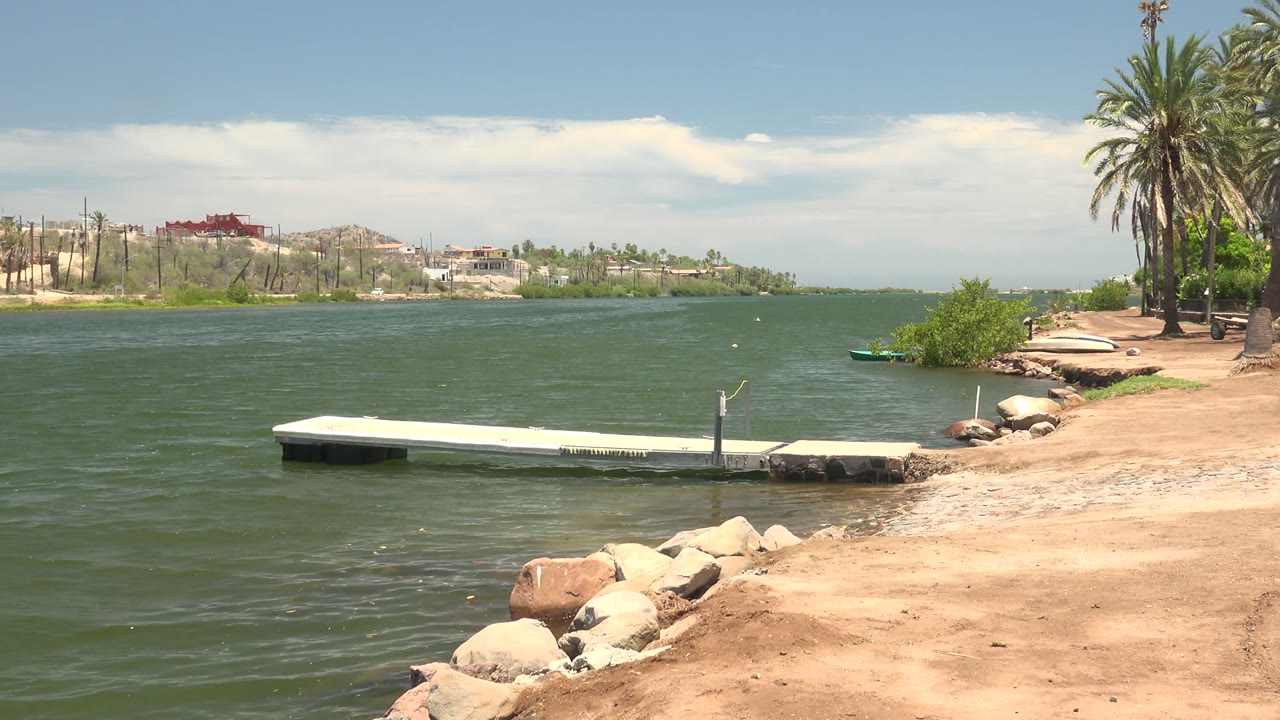 Opening frame of a Mulege riverbank with green water, a small white dock, rocks on the sandy edge, palms at right, distant buildings, and no visible people.