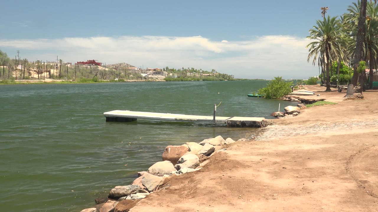 Mulege shoreline frame with more sandy bank visible at right, the low white dock over green water, foreground rocks, distant buildings, and no visible people.