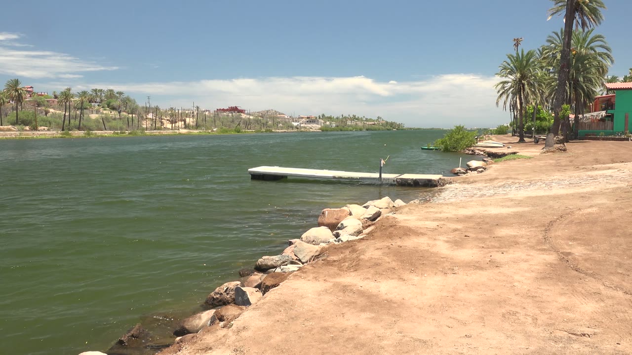Opening frame of a Mulege, Baja California Sur waterfront with green water, a white dock near the sandy rock-lined shore, palms at right, distant buildings across the water, and no visible people.