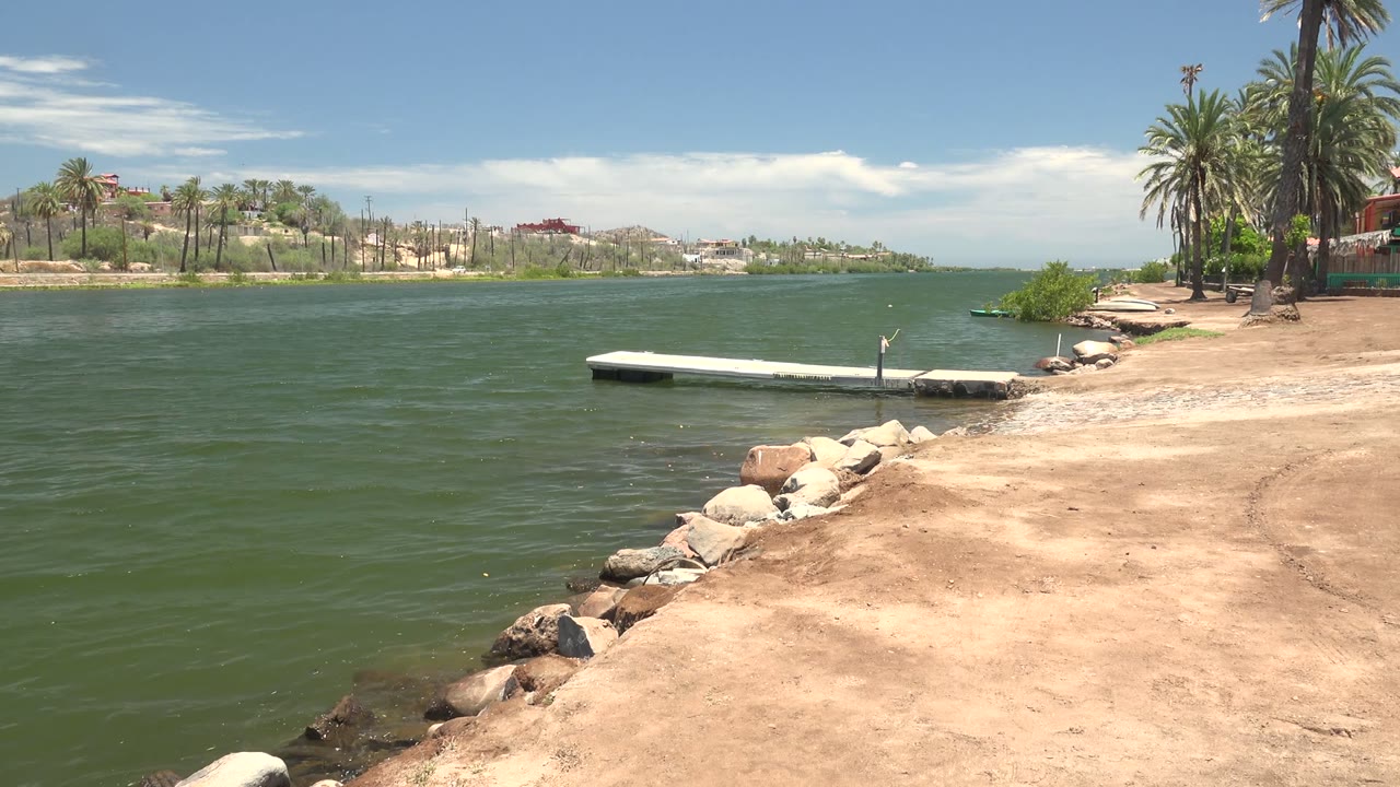 At 0:02, the Mulege shoreline remains framed from the sandy bank, with rocks leading toward the white dock, palms on the right edge, distant buildings across the water, and no visible people.