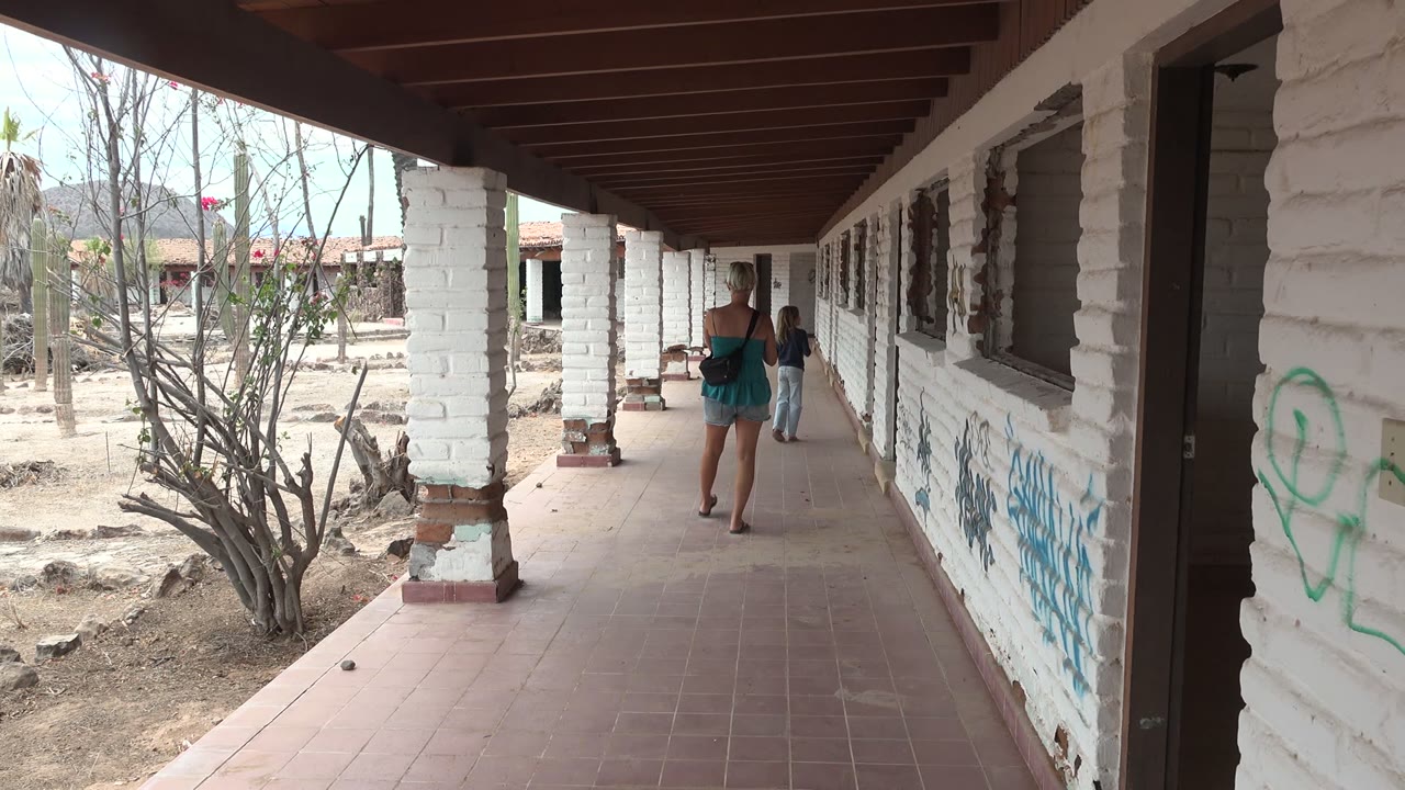 At 0:00 in Mulege, Baja California Sur, Mexico, two people walk away under a shaded corridor with white brick columns, tiled floor, graffiti, and dry vegetation outside.