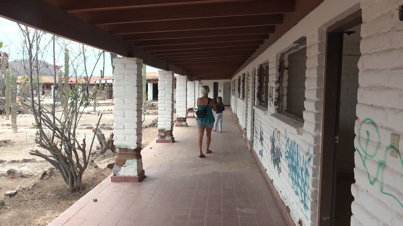 At 0:02 in Mulege, two people move deeper into the long porch while white columns divide the arid courtyard from the shaded tiled walkway.
