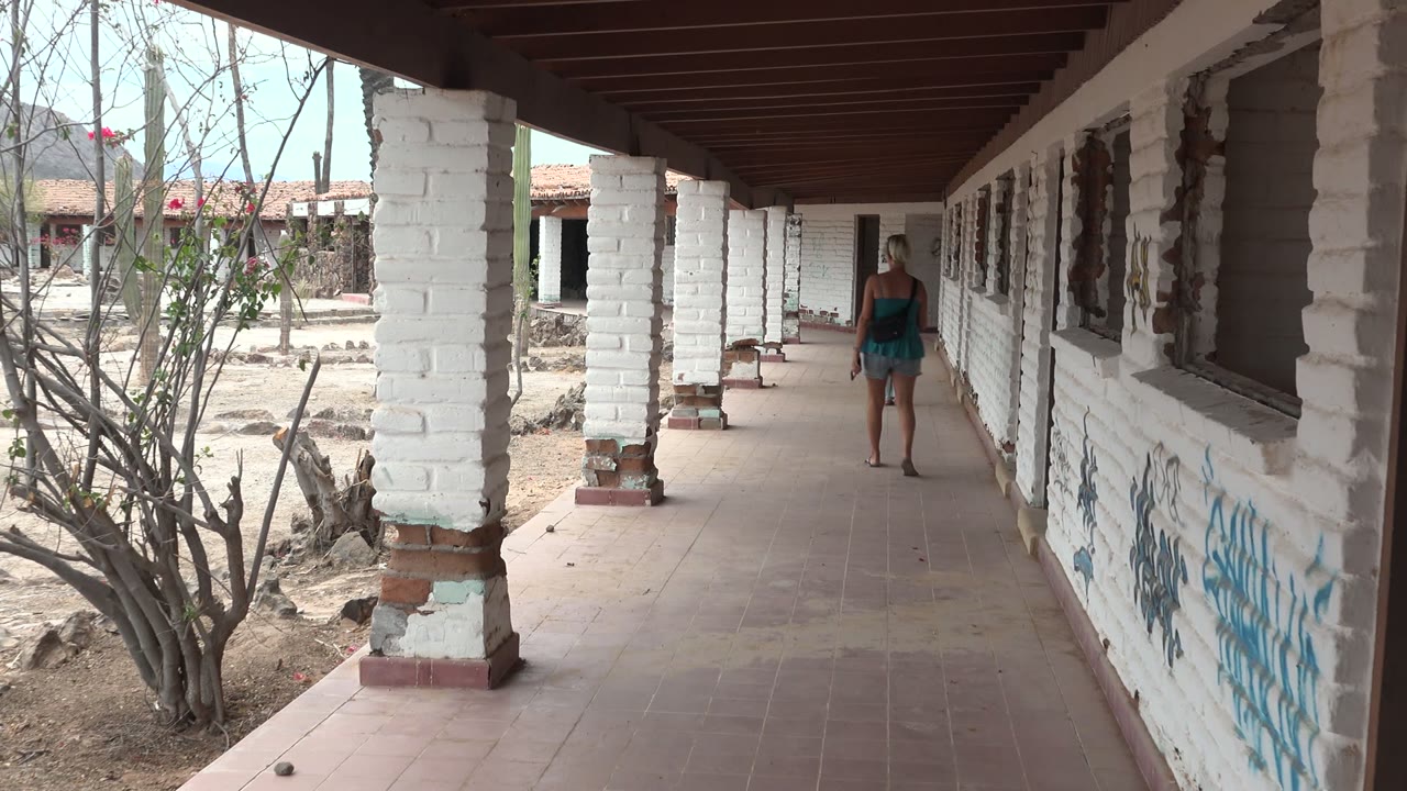 A person in a blue top walks away along a covered white brick corridor in Mulege, Mexico, with dry shrubs and low buildings visible outside.