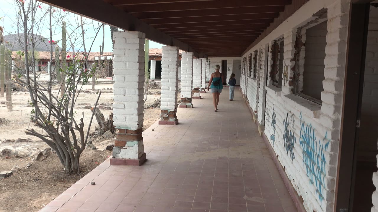 Two people are visible in the covered corridor in Mulege, Mexico, with the lead walker ahead and a second person farther back near the wall.