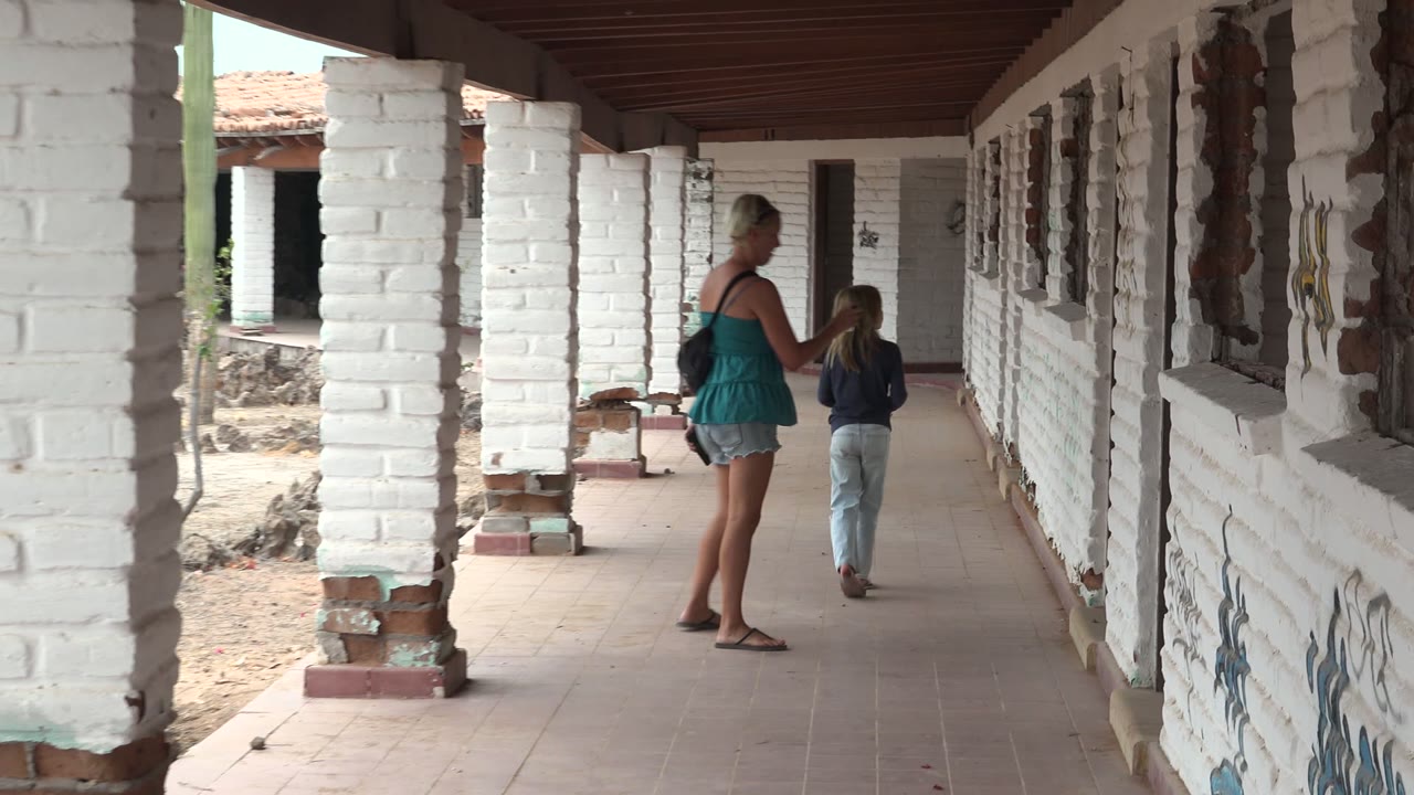 In Mulege, Mexico, a woman in a teal top walks behind a child along a shaded white brick corridor beside an open courtyard.