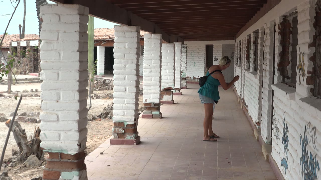 In Mulege, Mexico, the woman leans close to the corridor wall near rough window openings, with white columns and a dry courtyard visible to the left.