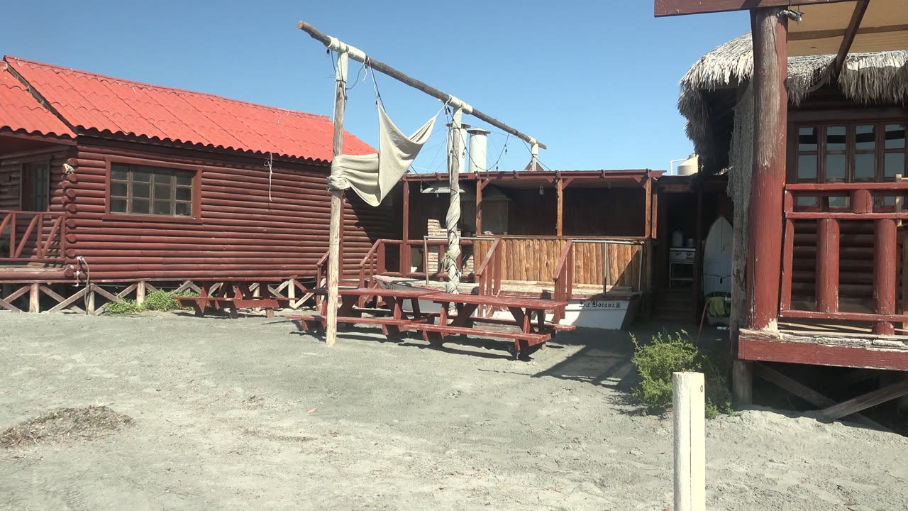 A red-roofed wooden beach cabin in La Bocana, Baja California Sur, with a hammock frame, picnic table, sandy foreground, and a partly visible person near the right doorway.