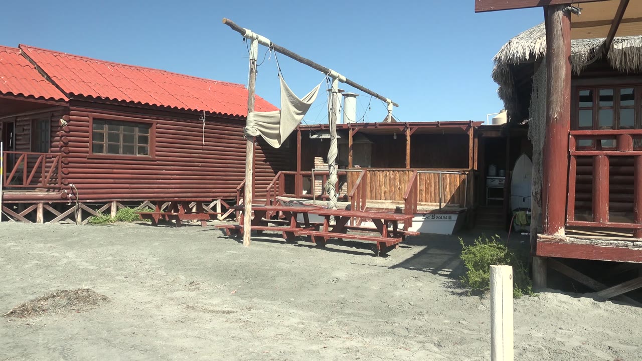 Wooden beach cabin with red roof, hammock frame, picnic table, and open doorway.